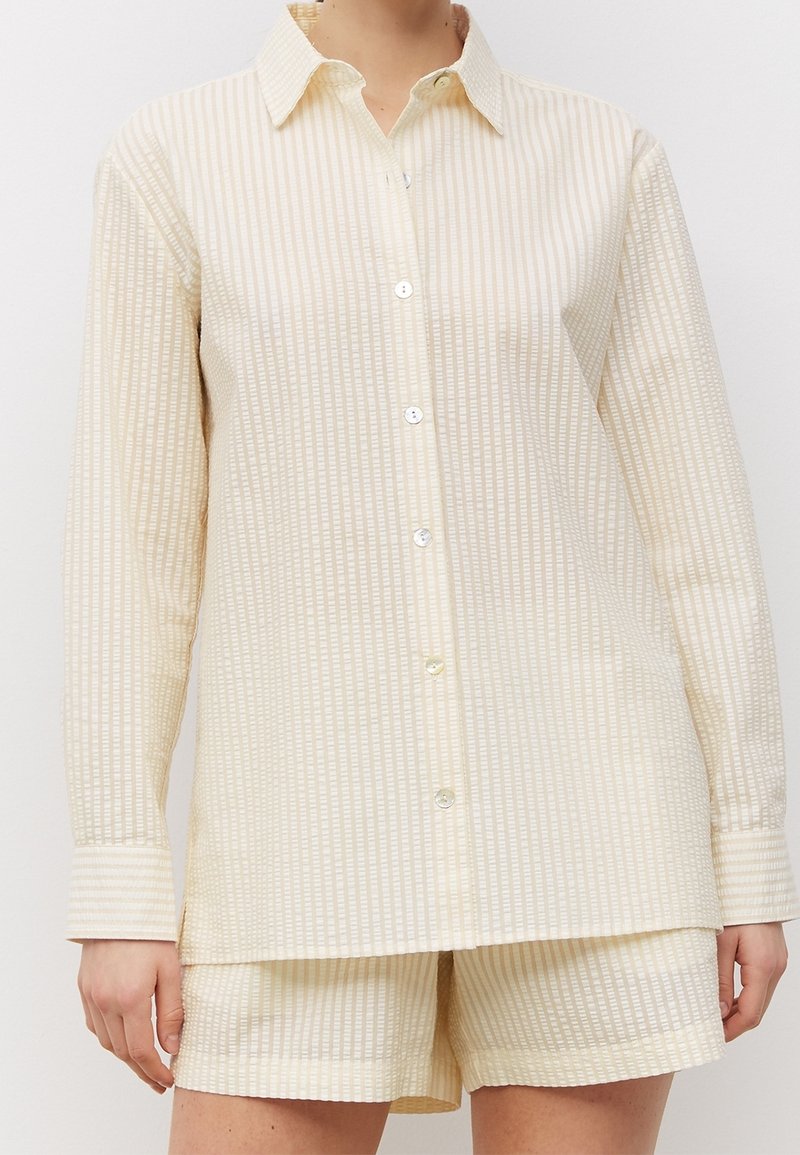 Femme portant une chemise à manches longues à rayures crème et blanches avec des boutons, accompagnée d'un short texturé assorti sur un fond uni.