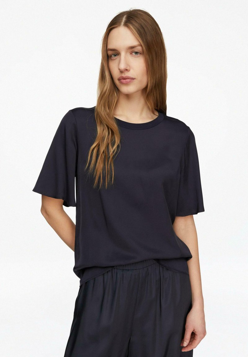 Junge Frau mit langen hellbraunen Haaren, die ein lockeres schwarzes Kurzarmoberteil und passende schwarze Hosen trägt, vor einem weißen Hintergrund.