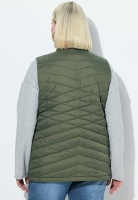 Olivgrüner, wattierter Weste mit Zickzack-Steppung, kombiniert mit einem grauen Langarmoberteil. Sie hat einen hohen Kragen und ein ärmelloses Design.