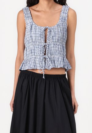 Vrouw draagt een blauw-wit geruit mouwloos cropped top met knopen aan de voorkant, gecombineerd met een zwarte, hooggetailleerde geplooide rok.