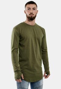 Olive zaļa garām piedurknēm t-krekls, izgatavots no mīksta auduma, ar apaļu apkakli un pieguļošām piedurknēm, saskaņots ar džinsiem, kuriem ir viegli dilti detaļas.