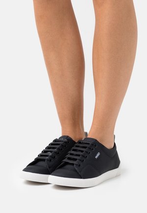 Schwarze Leinensneaker mit weißen Gummisohlen. Merkmale sind sechs Ösen, schwarze Schnürsenkel und ein Logo in der Nähe der Schnürsenkel. Minimalistisches Design.