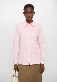 Camicia a maniche lunghe a righe rosa con ricamo di margherite bianche e colletto classico. Abbinata a una gonna khaki e una borsa di colore chiaro.