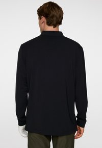 Mann in einem langen schwarzen Poloshirt und olivgrünen Hose, der mit dem Rücken zu einer schlichten Hintergrund steht.