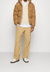Brown padded jacket, cream v-neck sweater, white button-up shirt with stripes, beige trousers, and light shoes with dark soles.