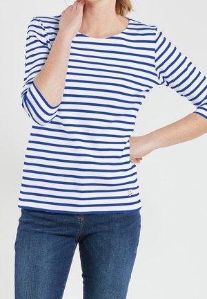 Femme portant un chemisier à manches trois-quarts blanc avec des rayures horizontales bleues et un jean bleu, une main sur la hanche, l'autre touchant le cou.