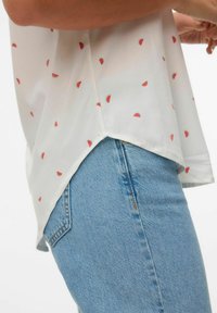 Hvid bluse med små print af vandmelonskiver, med korte ærmer og en længere bagkant, parret med lyseblå denimjeans.