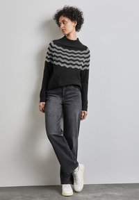 Schwarzer Strickpullover mit grauen Wellenmustern, hoher Kragen, kombiniert mit dunkelgrauen Jeans und weißen Sneakers, vor einem neutralen Hintergrund.