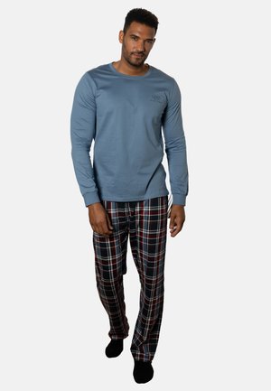 Mann trägt blaues Langarmhemd und rot-schwarz-weiß karierte Schlafanzughose, steht vor schlichtem hellem Hintergrund.