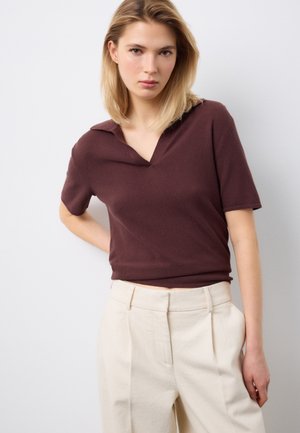 Giovane donna con capelli biondi che indossa un top marrone a maniche corte in maglia e pantaloni beige a vita alta, in piedi contro uno sfondo chiaro e semplice.