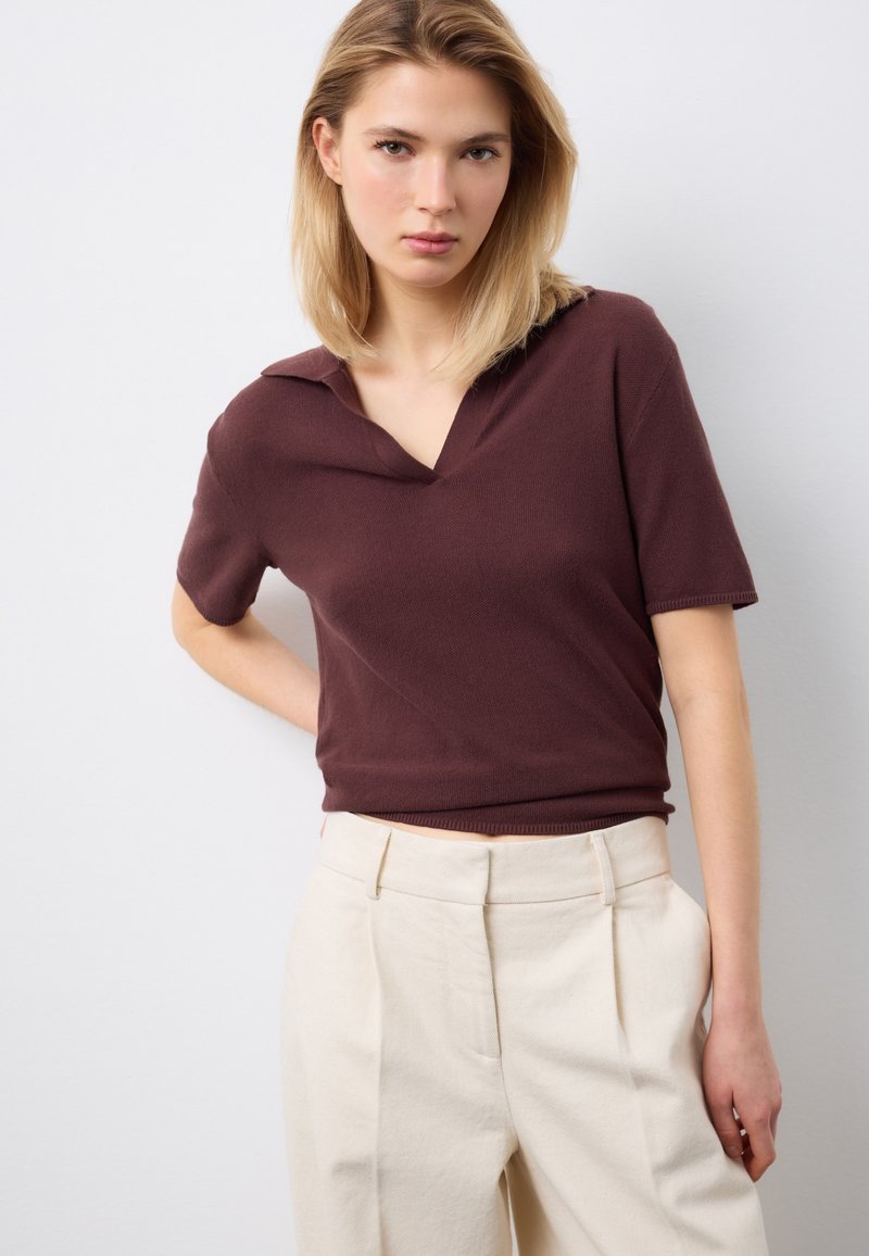 Junge Frau mit blondem Haar, die ein braunes, kurzärmeliges Strickoberteil und eine beige Hose mit hohem Bund trägt, vor einem schlichten hellen Hintergrund stehend.