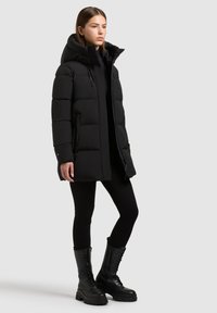 Cappotto imbottito nero con un grande cappuccio, chiusura con cerniera frontale e tasche laterali. Indossato sopra leggings neri e robusti stivali neri stringati.