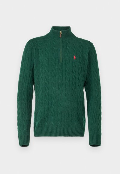 Žalias megztinis su aukštu apykakle, su ketvirčio užtrauktuku, su kabelių mezgimo raštu ir maža raudona logotipu ant krūtinės.