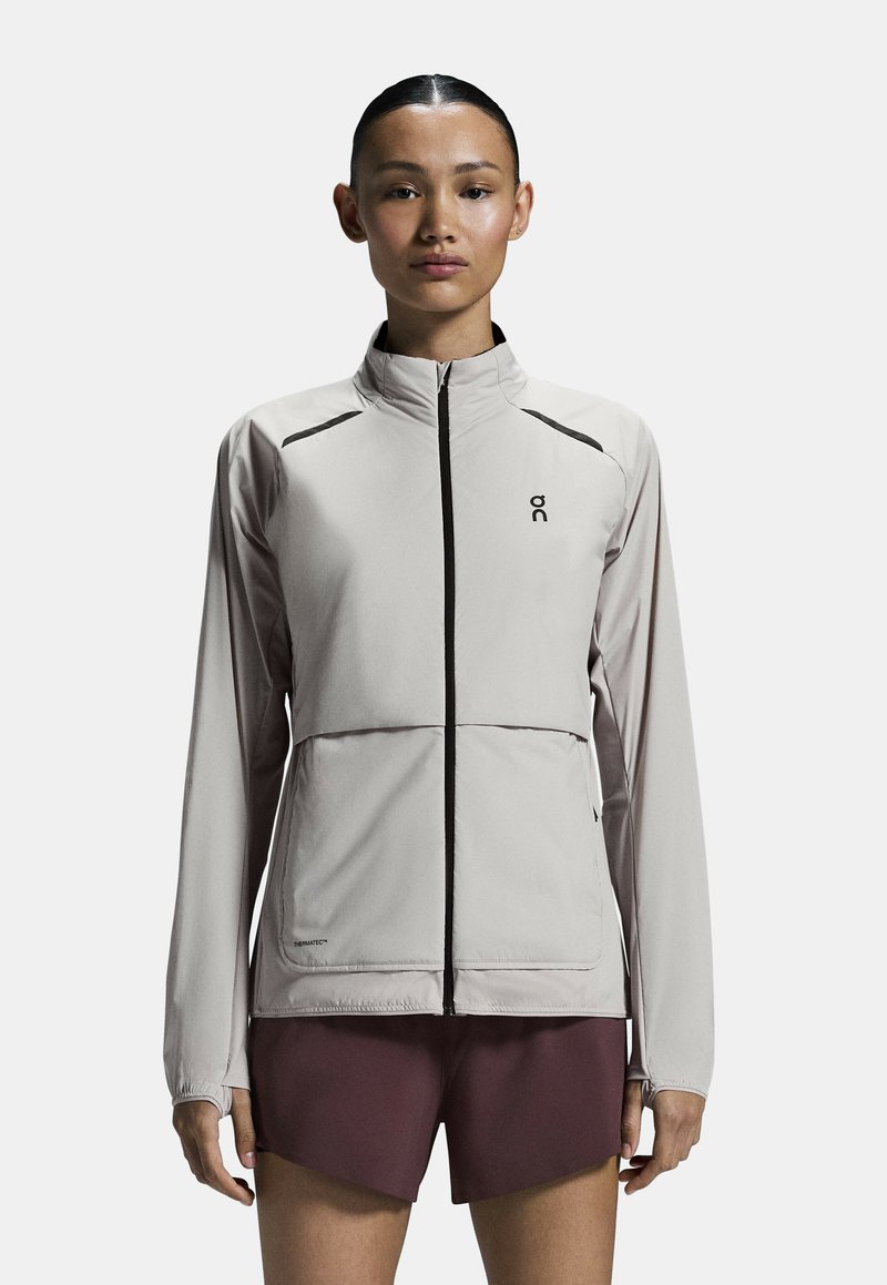 Chaqueta deportiva gris claro con un alto cuello, cremallera frontal completa y acentos negros en contraste. Presenta mangas largas y bolsillos frontales.