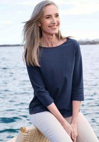 Marineblaues, leichtes Langarmoberteil mit weitem Ausschnitt, getragen mit hellbeigen Hosen, sitzend auf einer geflochtenen Oberfläche am Wasser.