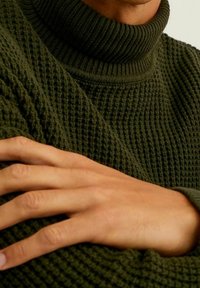 Pull col roulé vert olive en maille texturée avec un col haut. Gros plan sur les manches et une main reposant sur le tissu.
