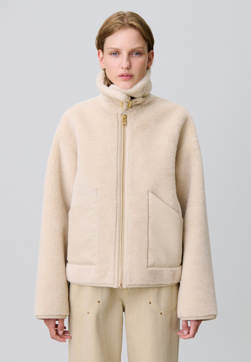 Giacca in pile beige con colletto alto, zip frontale e due ampie tasche a toppa. Caratterizzata da una consistenza morbida e una vestibilità comoda.