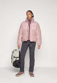 Woman standing with light pink padded jacket, dark pants, mauve sneakers, holding light gray and black backpack against plain background.