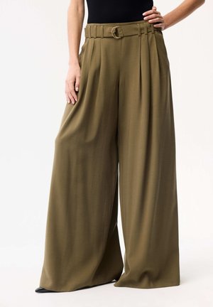 Kobieta nosząca oliwkowozielone spodnie z wysokim stanem i szerokimi nogawkami, z dopasowanym paskiem z okrągłą, złotą klamrą, w połączeniu z czarnymi butami.