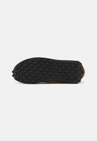 Semelle de chaussure en caoutchouc noire avec des motifs rectangulaires et des clous circulaires. dessus en tissu marron visible sur le bord. Texturé pour la prise et la traction.