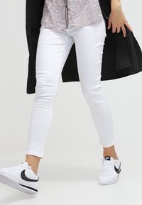 Bílé úzké džíny v kombinaci s blůzou s jemnými růžovými puntíky, černým kabátem a bílými teniskami s černým logem. Hladká látka, přiléhavý střih.