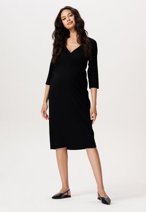 Schwarzes Stillkleid aus elastischem Stoff, mit Wickeldesign, Dreiviertelärmeln und knielangem Saum. Trägt schwarze Ballerinas.