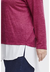 Långärmad topp i mörkrosa strukturerat material, lager på lager över en vit blus med rundad fåll, tillsammans med mörkblå jeans.