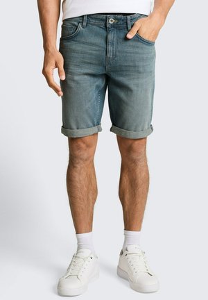 Mann trägt hochgekrempelte blaue Jeans-Shorts, weiße Socken und weiße Sneakers und steht mit einer Hand in der Tasche vor einfachem Hintergrund.