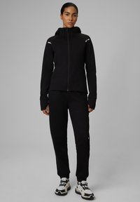 Femme debout en sweat à capuche noir zippé et pantalon de jogging avec des chaussures de sport blanches et noires sur fond gris.