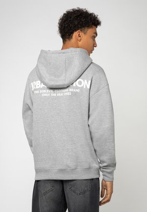 Grauer Hoodie mit weicher Textur, ausgestattet mit einer geräumigen Kapuze und oversized Passform. Auf dem Rücken befindet sich großer weißer Text mit dem Markennamen und dem Slogan.