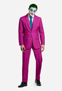 Heller pinker Anzug, passende Hose, kombiniert mit einer türkisfarbenen Krawatte. Gesicht weiß bemalt mit blauen Diamantmustern und roten Lippen. Schwarze Schuhe.