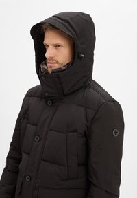 Man in een zwarte gewatteerde winterjas met capuchon en bontgevoerde kraag die naar links kijkt, op een effen witte achtergrond.