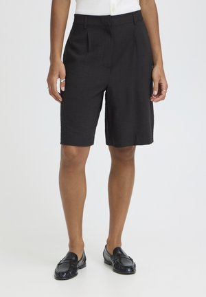 Osoba oblečená v čiernych, na mieru šitých šortkách po kolená, bielej tielkovej blúzke bez rukávov, čiernych mokasínach a so striebornými prsteňmi na prstoch, stojaca na jednoliatom bielom pozadí.