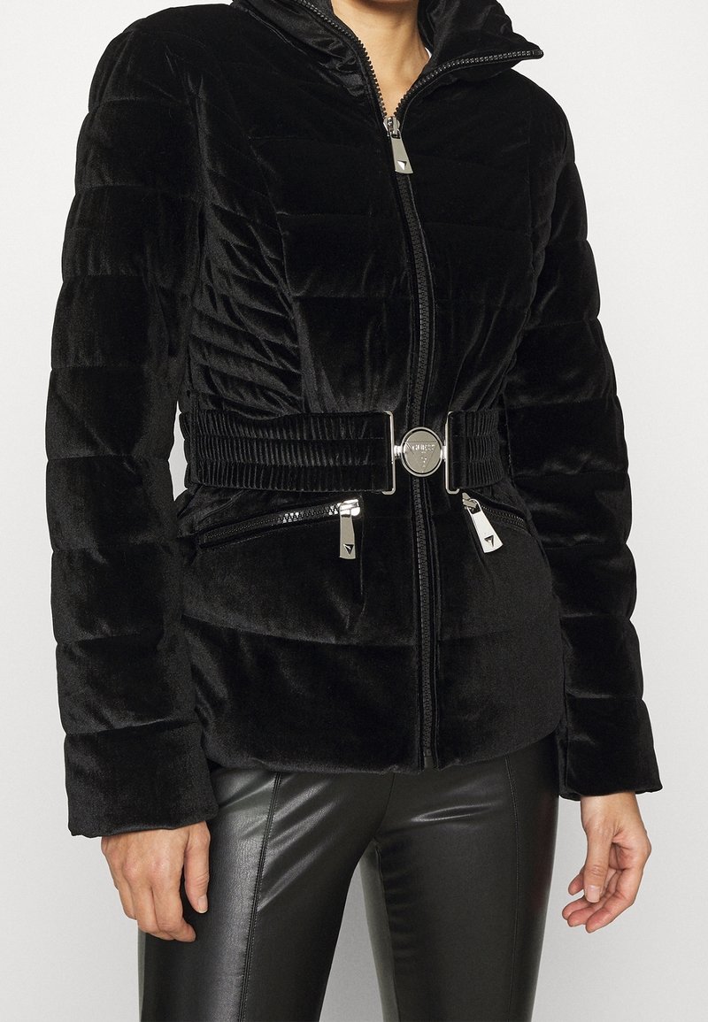 Veste matelassée noire avec col haut, taille ceinturée, poches zippées, portée par une personne portant un pantalon en cuir noir, debout devant un fond uni.