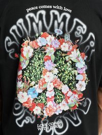 Sort t-shirt med et blomster-design af et fredstegn. Teksten siger "fred kommer med kærlighed" og "kærlighed kommer med fred." Livlige blomsterdetaljer.