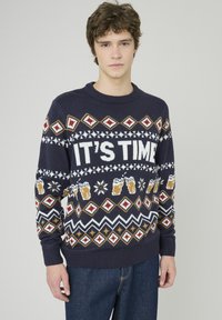 Pull en tricot bleu marine avec un design festif comportant l'inscription blanche "C'EST L'HEURE", des motifs de bière et des motifs géométriques en rouge et or.