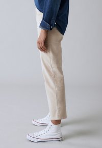Pantalones de pana beige con corte recto, bajos enrollados, combinados con zapatillas altas blancas y una camisa de mezclilla azul visible desde el lado.