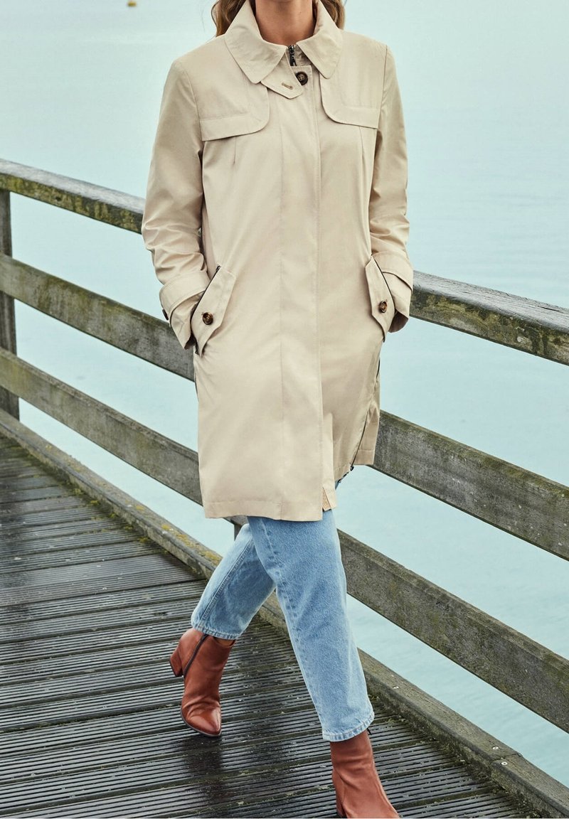 Personne portant un trench beige, un jean bleu et des bottines marron marchant sur une passerelle en bois au bord de l'eau.