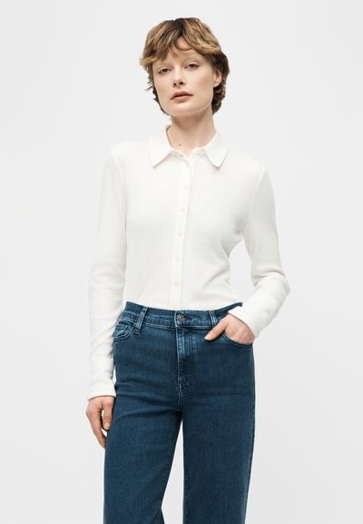 Γυναίκα με κοντά μαλλιά που φοράει λευκό πουκάμισο με μακριά μανίκια και κουμπιά και μπλε ψηλόμεσο τζιν, πόζαροντας με το ένα χέρι στην τσέπη.