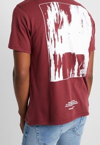Mørkerød bomulds t-shirt med et hvidt abstrakt grafik på ryggen. Har korte ærmer og en lige kant. Tekst nederst.