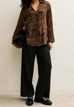 Vrouw die een bruine blouse met luipaardprint draagt, brede zwarte broek, zwarte loafers, en een opgevouwen zwarte handtas vasthoudt tegen een effen achtergrond.