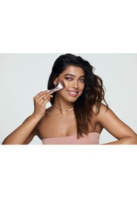 Femme aux longs cheveux foncés souriante, appliquant du maquillage avec un pinceau sur sa joue, portant un haut rose sans bretelles et des bijoux en or.