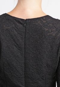 Vestido negro de encaje con un patrón texturizado, escote redondo y cierre de cremallera en la parte posterior. La tela parece ligera y semi-translúcida.