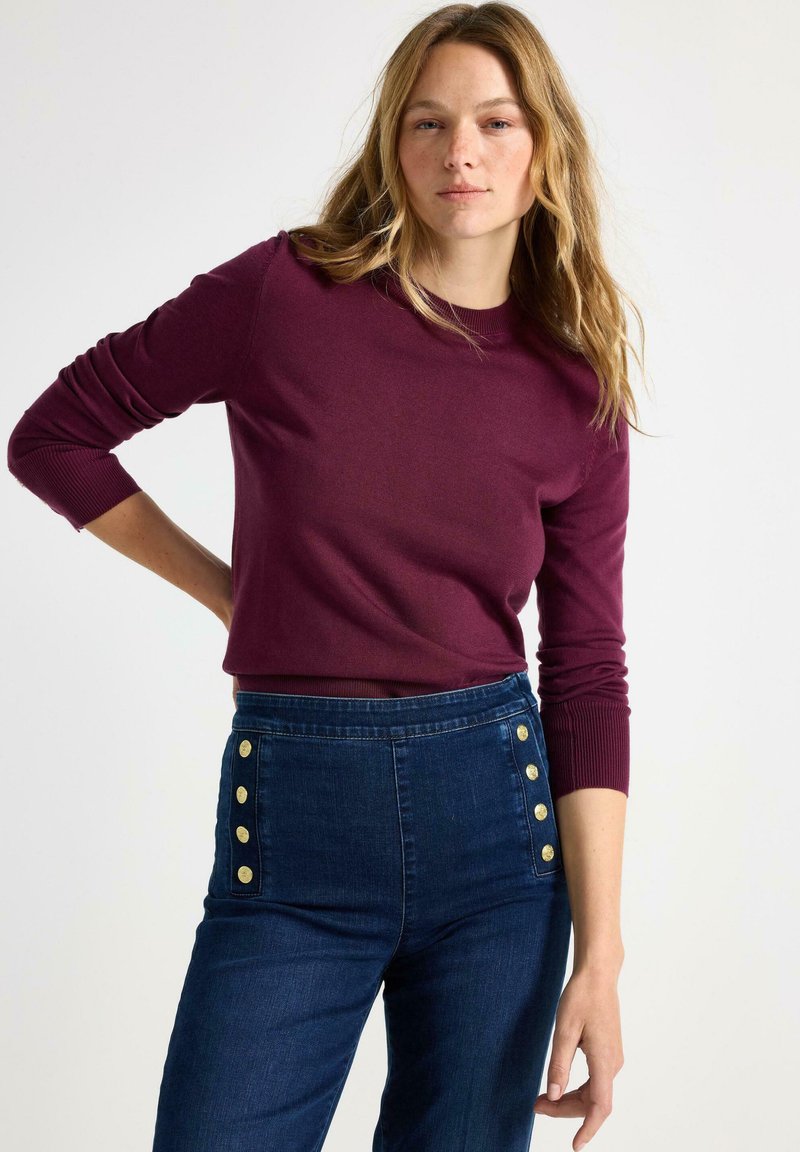 Vrouw met lang blond haar, gekleed in een wijnrode lange mouw trui en hoge taille donkerblauwe jeans met gouden knopen.