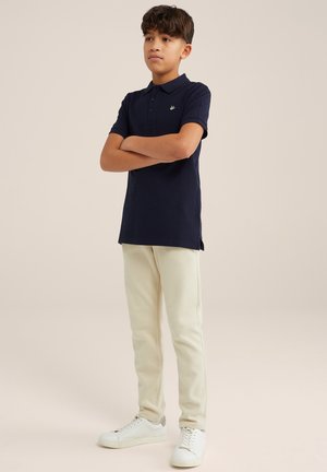Camisa polo navy con un pequeño logo, combinada con pantalones de chándal color crema y zapatillas blancas. Tejido suave, corte casual, mangas cortas.