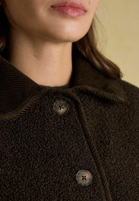 Hnedý chlpatej kabát s rebrovaným golierom, ktorý má dva okrúhle gombíky so zlatými detailami a textúrovanú látku.