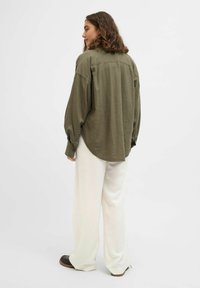 Chemise à manches longues vert olive avec un ourlet arrondi, coupe décontractée et poignets boutonnés, associée à un pantalon large crème et des chaussures noires.