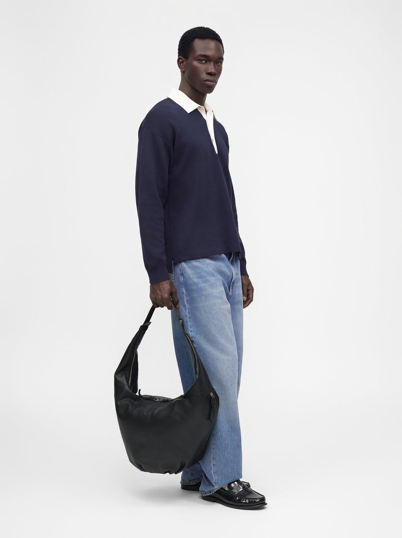 Un homme pose avec un maillot de rugby bleu marine, un jean bleu clair et des mocassins noirs. Il porte un grand sac noir en bandoulière sur un fond blanc uni.