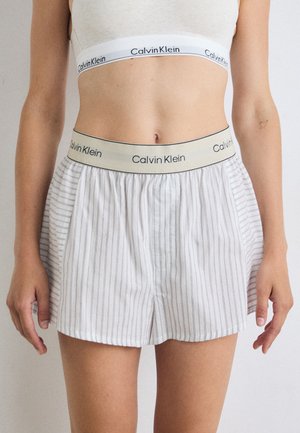 Modèle portant un crop top gris clair et des boxers rayés Calvin Klein, avec des ceintures visibles arborant le logo de la marque sur un fond neutre.
