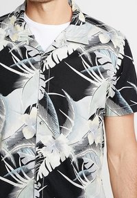Camisa negra de manga corta con un patrón de plantas tropicales y flores en azul claro y crema, con cuello estándar y botones.